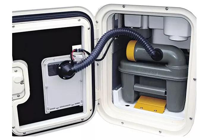 Offene wei&szlig;e Klappe eines eingebauten Fachs; darin ein grauer Kunststoffbeh&auml;lter mit gelber Abdeckung und ein schwarzer flexibler Schlauch, links an einem runden Anschluss befestigt; wei&szlig;e T&uuml;r mit schwarzem Innenteil und rechteckiger wei&szlig;er Platte
