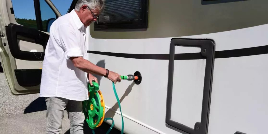 Person schlie&szlig;t gr&uuml;nen Wasserschlauch mit Aufroller an einen Anschluss an der Seite eines wei&szlig;en Wohnmobils; offene T&uuml;r links