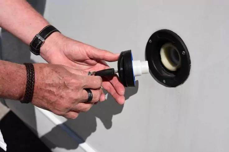 Zwei H&auml;nde mit Ringen, Armbanduhr und Armband halten ein rundes schwarzes Teil und einen schwarzen Stecker vor einer wei&szlig;en Wand mit rundem eingelassenem Einsatz