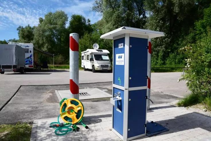 Blaue Versorgungss&auml;ule mit Anschl&uuml;ssen, gr&uuml;n-gelbe Schlauchtrommel auf dem Boden, wei&szlig;es Wohnmobil im Hintergrund, rot-wei&szlig;e Pfosten, Asphaltfl&auml;che, B&auml;ume und blauer Himmel