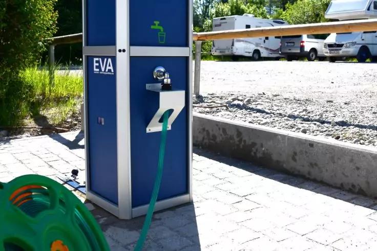 Blaue S&auml;ule mit der Aufschrift "EVA", Wasserhahn und gr&uuml;nem Schlauch, gr&uuml;ner Schlauchaufroller auf gepflastertem Boden, Wohnmobile und B&auml;ume im Hintergrund