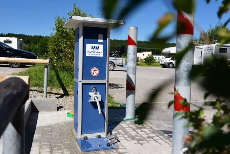 Blaue S&auml;ule mit Wasserhahn, Schlauchanschluss und Schild auf einem gepflasterten Platz; im Hintergrund Wohnmobile, Pflanzen und rot-wei&szlig;e Pfosten