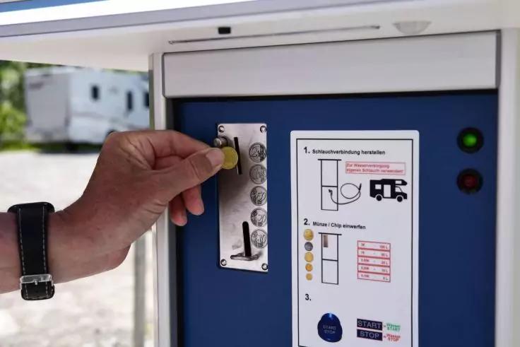 Hand mit schwarzer Armbanduhr f&uuml;hrt eine M&uuml;nze in einen Metallschlitz an einem blauen Bedienfeld, daneben Anleitungsaufkleber und zwei Kontrollleuchten; unscharfer Wohnwagen im Hintergrund