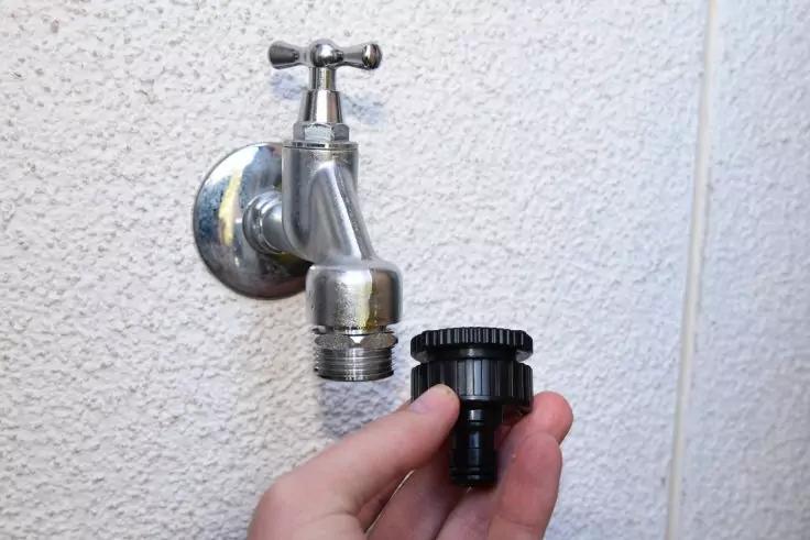 Eine Hand h&auml;lt einen schwarzen Kunststoffadapter vor einem metallenen Wandwasserhahn an einer wei&szlig;en strukturierten Wand