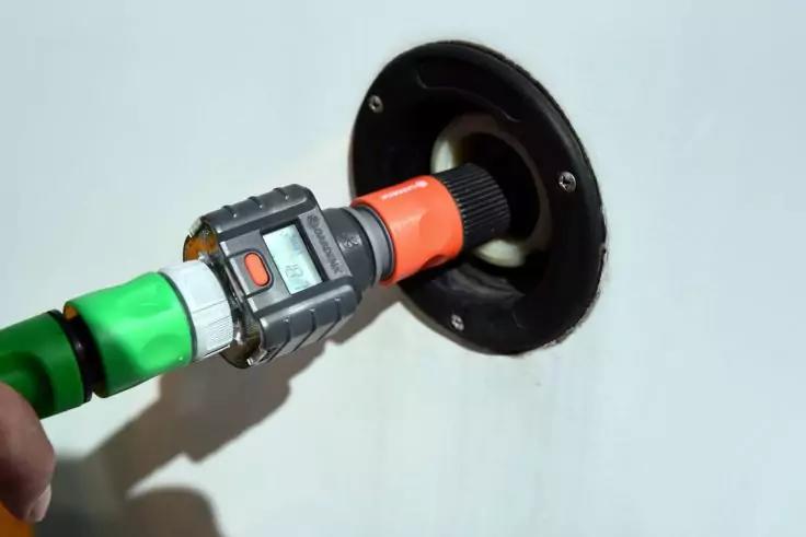 Hand h&auml;lt einen grau-schwarzen Steckverbinder mit orangefarbener H&uuml;lse, der in eine runde schwarze Wandbuchse eingef&uuml;hrt wird; ein gr&uuml;nes Kabel ist links sichtbar