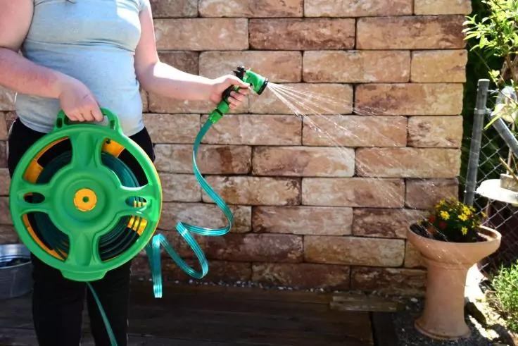 Person h&auml;lt gr&uuml;ne Gartenschlauchrolle in der linken Hand und Spr&uuml;hd&uuml;se in der rechten Hand, Wasserstrahl sichtbar, vor einer Sandsteinmauer; rechts ein Topf mit gelben Blumen