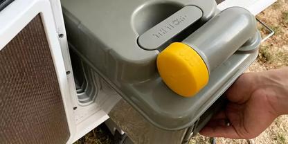 Hand h&auml;lt grauen Kunststoffbeh&auml;lter mit rundem gelben Verschluss neben einer wei&szlig;en &Ouml;ffnung mit L&uuml;ftungsgitter