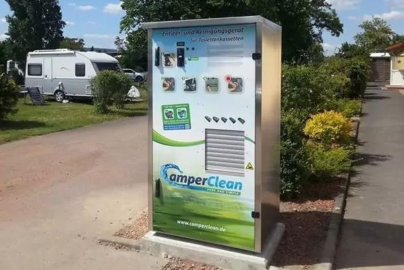 Metallene Säule mit Aufschrift 'CamperClean', Bedienfeld und Piktogrammen auf einem Campingplatz; daneben Wohnwagen, Rasen, Bäume und blauer Himmel