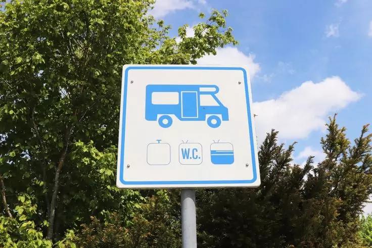 Quadratisches Schild mit blauem Wohnmobil-Piktogramm und drei Symbolen darunter: Wassersymbol, Quadrat mit 'W.C.' und Behältersymbol; Schild auf einem Pfosten vor Bäumen und blauem Himmel