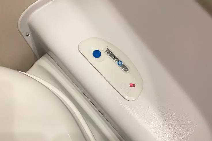 Weiße Toilette; ovales Bedienfeld mit blauem Knopf und kleinem roten Aufkleber auf der Oberseite des Spülkastens