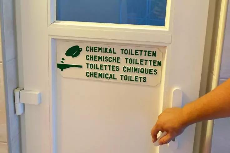 Weiße Tür mit kleinem Schild 'CHEMIKAL TOILETTEN / CHEMISCHE TOILETTEN / TOILETTES CHIMIQUES / CHEMICAL TOILETS' und einer Hand am Türgriff