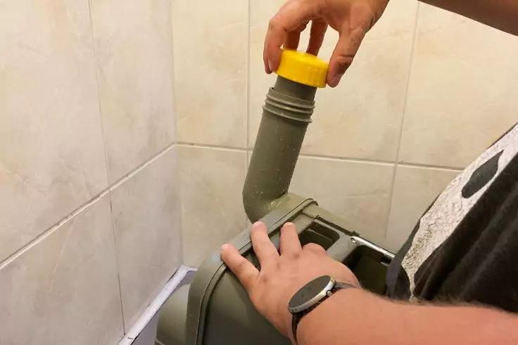 Hände einer Person halten ein grünes, gebogenes Kunststoffrohr mit gelbem Deckel über einem grünen Behälter; geflieste Wand im Hintergrund