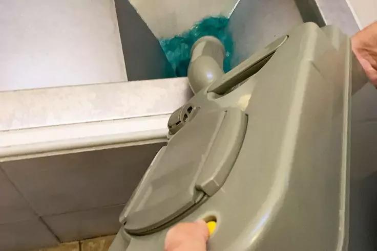 Zwei Hände halten einen grauen Kunststoffbehälter mit aufgeklapptem Deckel, der über eine größere Öffnung gekippt wird; im hinteren Behälter ist ein blau-grüner, faseriger Gegenstand sichtbar