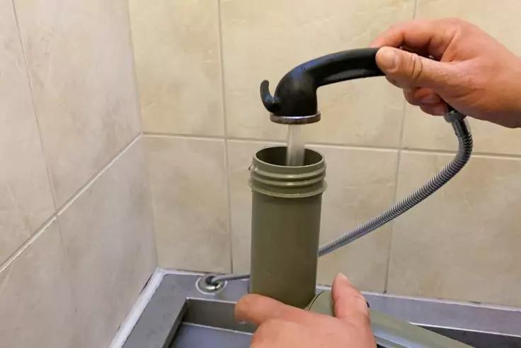 Hände halten einen schwarzen Brausekopf an einem flexiblen Schlauch über einem grünen zylindrischen Behälter; Wasser fließt in ein Edelstahlspülbecken, geflieste Wand im Hintergrund