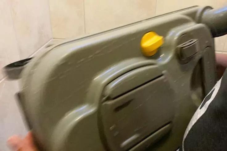 Nahaufnahme eines olivgrünen Kunststoffgehäuses mit gelbem Knopf, seitlichem Griff und sichtbaren Verschlüssen; Hände und schwarzes Kleidungsstück am rechten Rand; helle Wandfliesen im Hintergrund