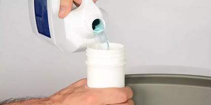 Hand gie&szlig;t blaue Fl&uuml;ssigkeit aus einer Kunststoffflasche in ein offenes wei&szlig;es Gef&auml;&szlig; &uuml;ber einer grauen Fl&auml;che