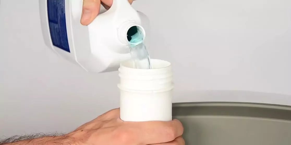 Hand gie&szlig;t blaue Fl&uuml;ssigkeit aus einer Kunststoffflasche in ein offenes wei&szlig;es Gef&auml;&szlig; &uuml;ber einer grauen Fl&auml;che