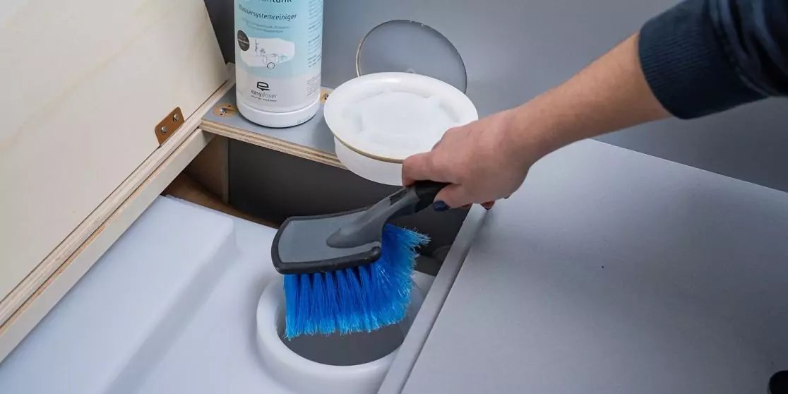 Hand mit blauem B&uuml;rstenkopf und grauem Handfeger &uuml;ber runder &Ouml;ffnung in wei&szlig;er Fl&auml;che; wei&szlig;er Beh&auml;lter und Dose im Hintergrund