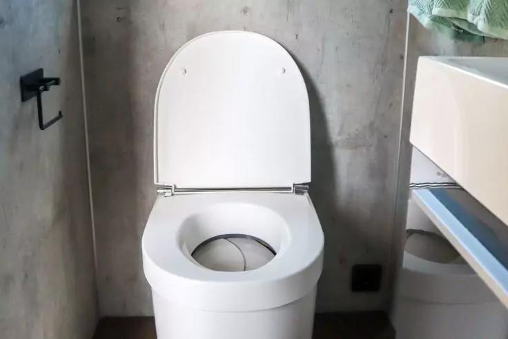 Wei&szlig;e Toilette mit ge&ouml;ffnetem Deckel vor grauer Wand; links schwarzer Toilettenpapierhalter; rechts wei&szlig;er Schrank mit Handtuch