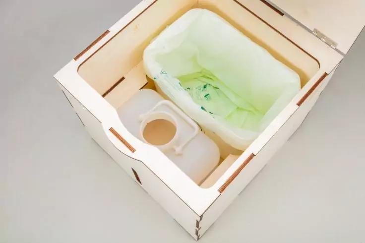 Heller rechteckiger Holzkasten mit Ausfr&auml;sungen, darin ein wei&szlig;er Kunststoffkanister mit Henkel und ein rechteckiger Kunststoffbeh&auml;lter mit hellgr&uuml;ner Plastikfolie ausgelegt