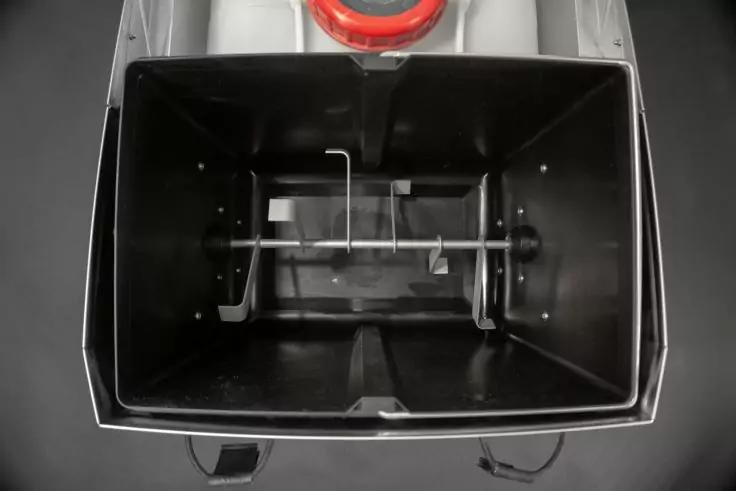 Blick von oben in einen rechteckigen schwarzen Beh&auml;lter mit einer quer laufenden Metallachse und einem gebogenen Metallhaken, am oberen Rand sichtbar ein rotes rundes Teil