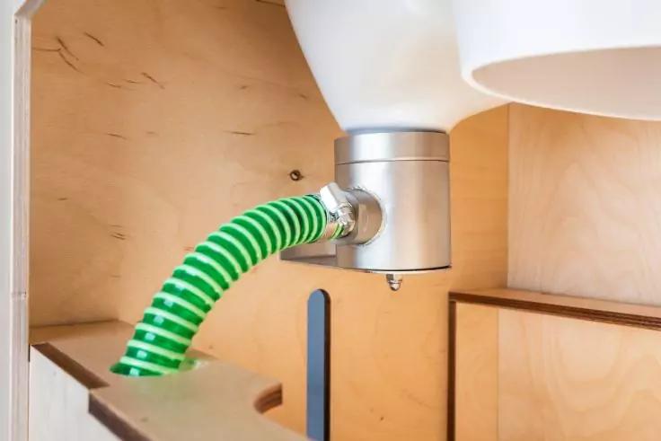 Innenraum eines Holzschranks mit wei&szlig;em runden Bauteil oben, silbernem Anschluss, an dem ein gr&uuml;n-wei&szlig; gestreifter Schlauch befestigt ist, und einem schwarzen senkrechten Stab