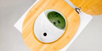 Aufsicht auf offenen hellen Holz-Toilettensitz mit wei&szlig;er Keramiksch&uuml;ssel, gr&uuml;nliche Fl&uuml;ssigkeit in der Sch&uuml;ssel, Toilettenpapier, Metallscharniere und grauer Boden