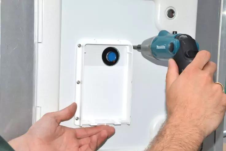 Eine Hand h&auml;lt eine wei&szlig;e Kunststoffabdeckung mit rundem Ausschnitt an einer wei&szlig;en Platte, eine andere Hand setzt einen blauen Akkuschrauber an einer Schraube an