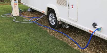 Seitenansicht eines Wohnwagens auf Gras- und Kiesfl&auml;che; Reifen sichtbar; blauer Schlauch ist an eine runde Einlass&ouml;ffnung an der Fahrzeugseite angeschlossen und f&uuml;hrt zu einem kleinen Versorgungspoller; grauer Schlauch und schwarzes Kabel liegen auf