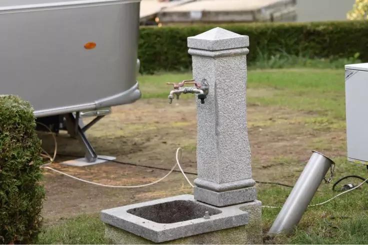 Graue steinerne S&auml;ule mit Metallhahn und rechtemcktem Becken davor; auf dem Boden liegende Metallstange, Wohnwagenst&uuml;tze links und Hecke im Hintergrund