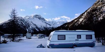 Wei&szlig;er Wohnwagen auf verschneitem Boden; im Hintergrund weitere Wohnwagen, Nadelb&auml;ume, schneebedeckte Berge und blauer Himmel mit Wolken