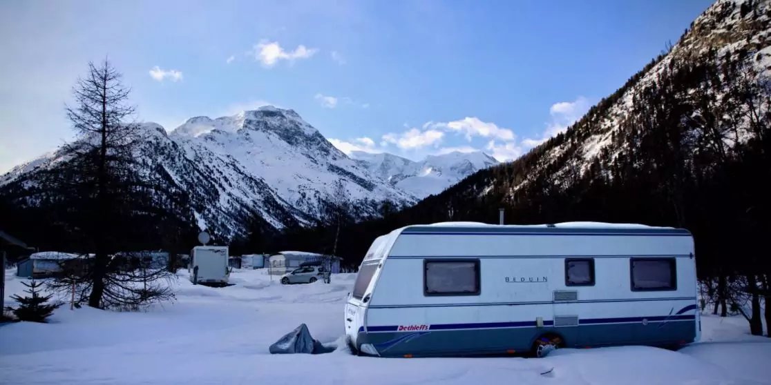 Wei&szlig;er Wohnwagen auf verschneitem Boden; im Hintergrund weitere Wohnwagen, Nadelb&auml;ume, schneebedeckte Berge und blauer Himmel mit Wolken