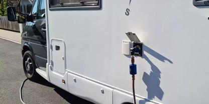 Seitenansicht eines wei&szlig;en Wohnmobils mit Vorderrad, ge&ouml;ffneter Au&szlig;enanschlussdose, blauem Stecker und Kabel, das auf den Boden f&uuml;hrt; Schatten an der Karosserie