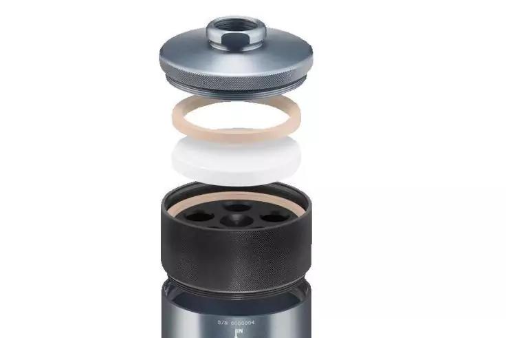 Auseinandergebaute zylindrische Metallteile auf wei&szlig;em Hintergrund: oberer silberner Deckel, beige Dichtung, wei&szlig;er Ring, schwarzes zylindrisches Teil mit drei runden &Ouml;ffnungen, unterer silberner Zylinder