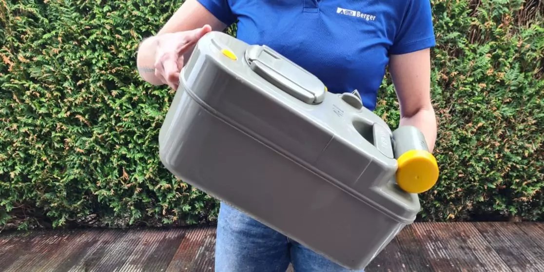 Person in blauem Shirt h&auml;lt einen grauen Kunststoffkanister mit gelben Verschl&uuml;ssen vor einer Hecke