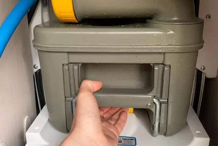 Hand greift Griff einer graugr&uuml;nen Kunststoffbox mit Metallstange, blaues Rohr links und wei&szlig;er Hintergrund