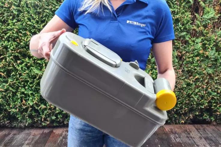 Person tr&auml;gt blaues Poloshirt und h&auml;lt einen grauen Kunststoffkanister mit gelbem Deckel vor einer Hecke