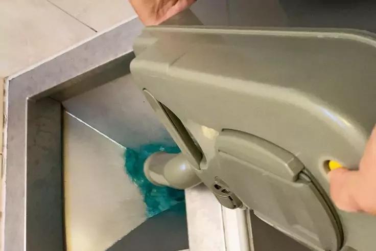 Zwei H&auml;nde halten ein graues Kunststoffteil mit gelbem Knopf &uuml;ber einer offenen Metall&ouml;ffnung; in der &Ouml;ffnung ist blauer Schaum sichtbar