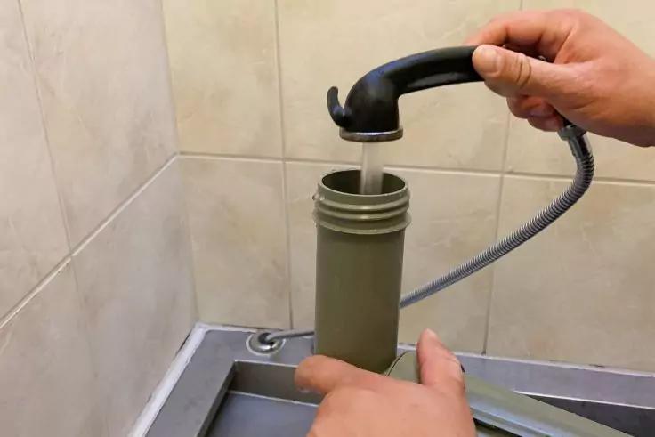 Zwei H&auml;nde halten eine schwarze Brause, Wasser flie&szlig;t in einen offenen gr&uuml;nen zylindrischen Beh&auml;lter; geflieste Wand und Sp&uuml;lbeckenrand sichtbar