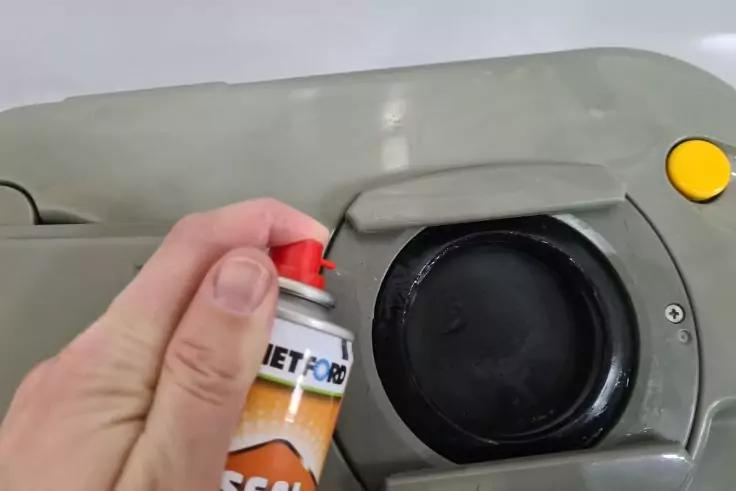 Hand h&auml;lt Spraydose neben runder schwarzer &Ouml;ffnung auf grauer Kunststofffl&auml;che mit gelbem Knopf