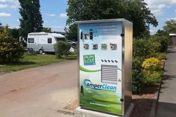 Metallene Stele mit Logo 'CamperClean' und Bedienelementen auf einem Campingplatz; Wohnwagen, B&auml;ume und Weg im Hintergrund