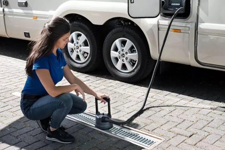 Frau kniet auf gepflastertem Boden neben einem wei&szlig;en Wohnmobil, h&auml;lt ein rundes schwarzes Ger&auml;t &uuml;ber einem Abflussgitter; ein schwarzes Kabel oder Schlauch verl&auml;uft zum Wohnmobil