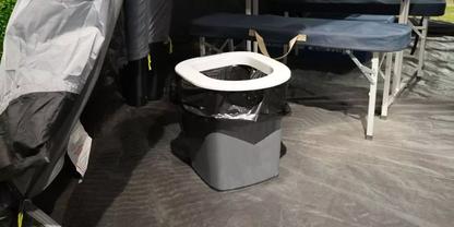 Grauer Kunststoffbeh&auml;lter mit wei&szlig;em ovalen Sitzring und schwarzer Plastikt&uuml;te, stehend auf dunkelgrauem Boden in einem Zelt; links eine graue Abdeckung, im Hintergrund zusammengeklappte blaue Plattformen mit Metallgestell