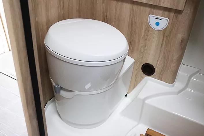 Wei&szlig;e kompakte Toilette mit geschlossenem Sitz auf zylindrischem Beh&auml;lter, neben einer holzverkleideten Wand mit rundem Loch und blauem Schalter auf wei&szlig;em Boden