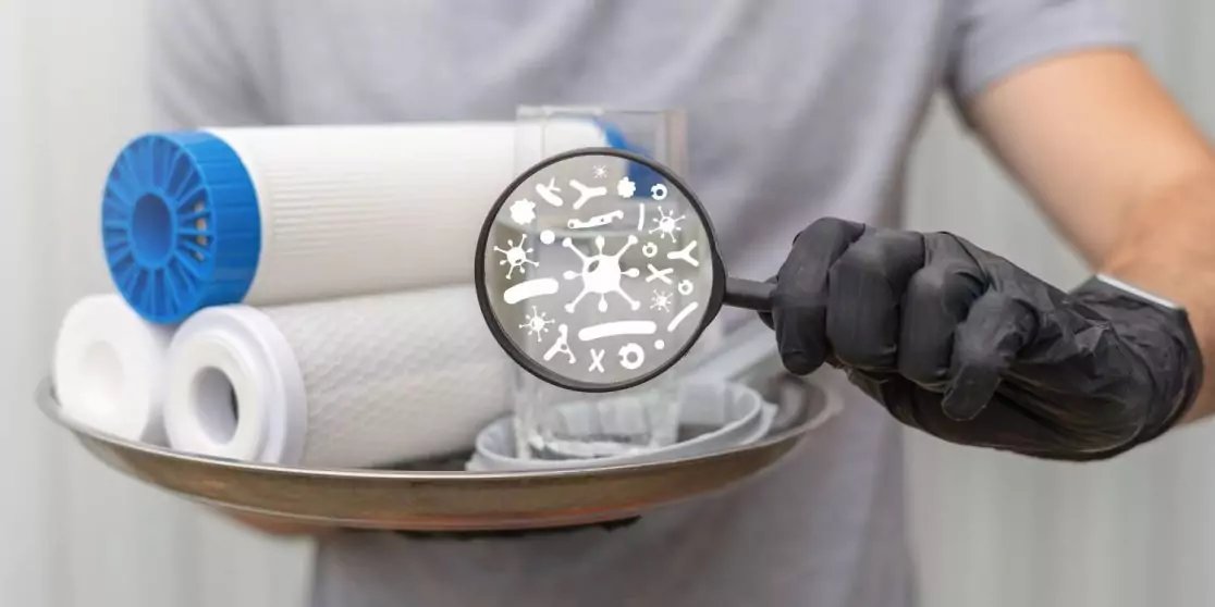 Tablett mit mehreren wei&szlig;en, zylindrischen Filterpatronen; eine Hand mit schwarzem Handschuh h&auml;lt eine Lupe &uuml;ber den Patronen, in der stilisierte Mikroben zu sehen sind