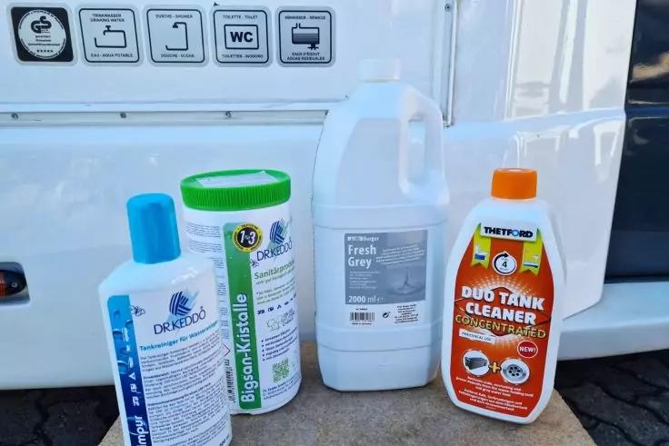 Quatre contenants en plastique avec &eacute;tiquettes de diff&eacute;rentes couleurs &ndash; &agrave; gauche : bouteille blanche avec bouchon bleu, ensuite : cylindre blanc avec couvercle vert, au centre : bidon blanc avec poign&eacute;e et &eacute;tiquette grise, &agrave; droite : bouteille blanche avec &eacute;tiquette orange ; en arri&egrave;re-plan : flanc d&rsquo;un v&eacute;hicule avec une rang&eacute;e de pictogrammes