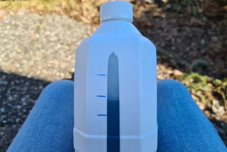 Wei&szlig;e Kunststoffflasche mit Schraubverschluss und vertikalem transparenten Messstreifen mit blauen Markierungen, liegt auf blauer Jeans; im Hintergrund Schotter und Gras