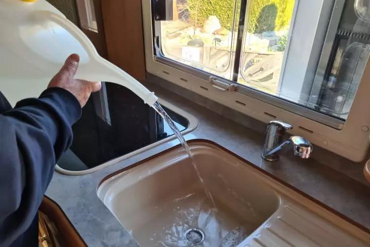 Hand gie&szlig;t Wasser aus einer wei&szlig;en Gie&szlig;kanne in ein Sp&uuml;lbecken unter einem Fenster