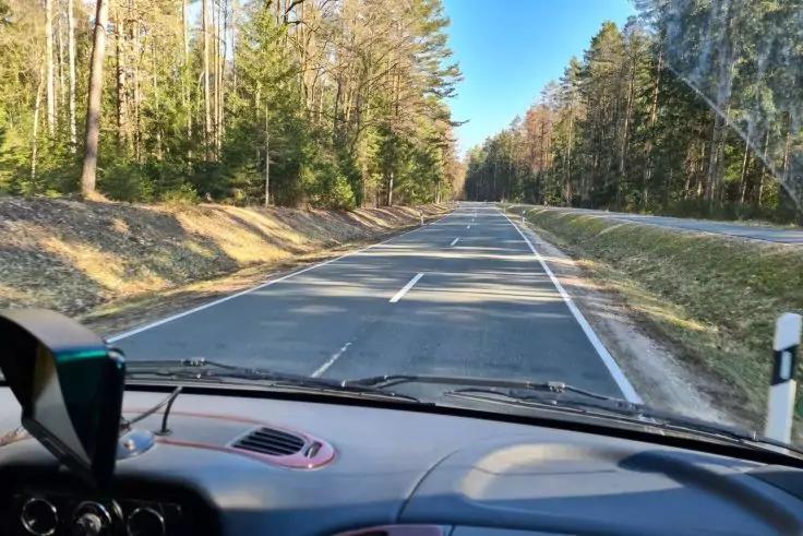 Blick aus einem Fahrzeug durch die Windschutzscheibe auf eine zweispurige Asphaltstra&szlig;e mit Mittel- und Randmarkierungen, B&auml;ume beidseits, Armaturenbrett und Scheibenwischer im Vordergrund