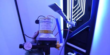 Hand mit Schutzhandschuh h&auml;lt ein zylindrisches Ger&auml;t mit Etikett; links ein runder Manometer; daran angeschlossene Schl&auml;uche und Kabel; im Hintergrund ein vertikales, beleuchtetes Paneel mit wei&szlig;en Linien und blauem Licht